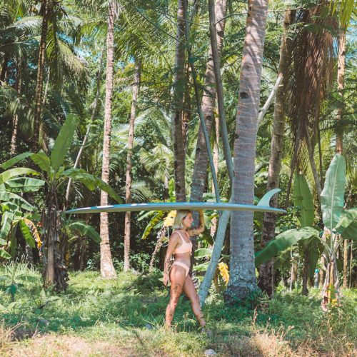 A woman carries a surfboard through a dense tropical jungle, surrounded by lush greenery.